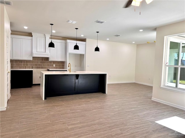 kitchen with ceiling fan, a kitchen island with sink, hanging light fixtures, decorative backsplash, and white cabinets