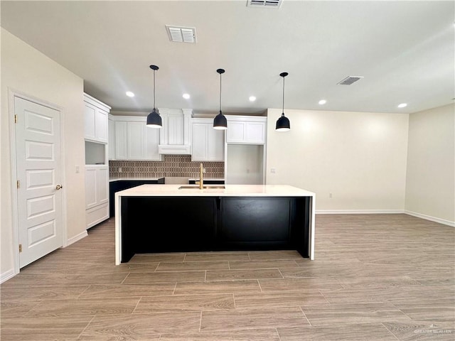 kitchen featuring backsplash, a kitchen island with sink, sink, hanging light fixtures, and white cabinetry