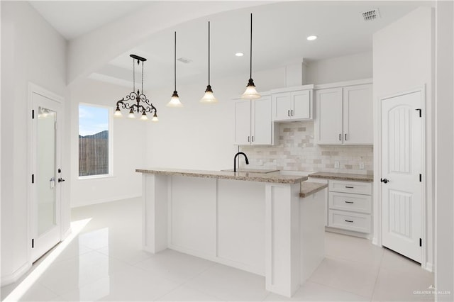 kitchen with a kitchen island with sink, light tile patterned floors, decorative light fixtures, light stone counters, and white cabinetry