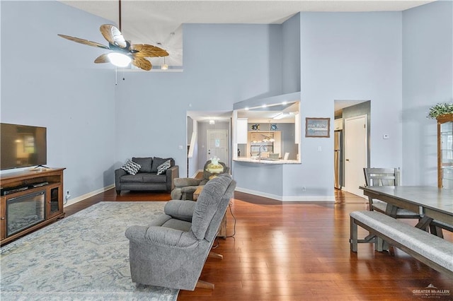 living room with a high ceiling, a ceiling fan, a glass covered fireplace, wood finished floors, and baseboards