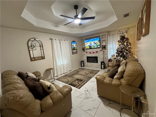 living room with a raised ceiling, ceiling fan, and a stone fireplace