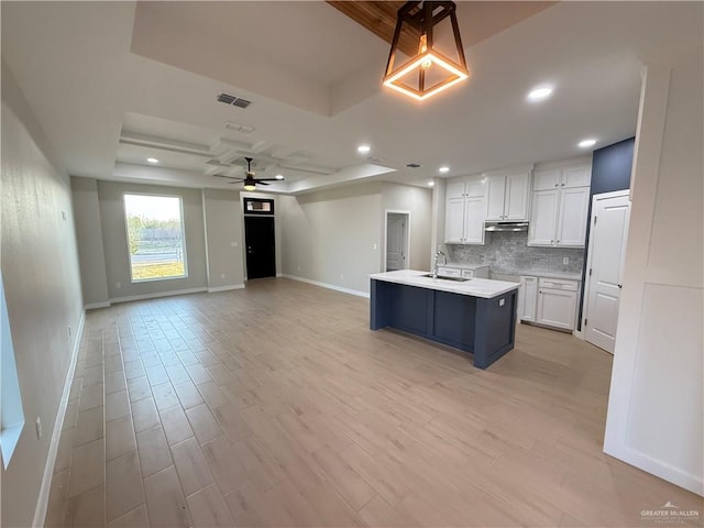 kitchen with white cabinetry, a kitchen island with sink, backsplash, and a tray ceiling