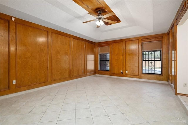 empty room with light tile patterned floors, a raised ceiling, a ceiling fan, wood walls, and baseboards