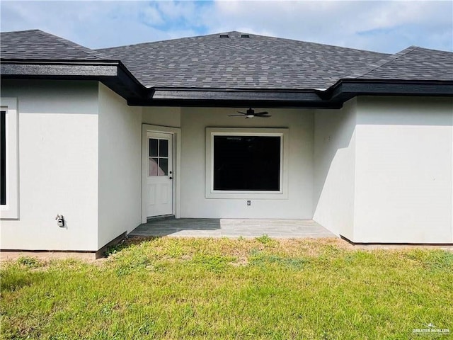 property entrance featuring a ceiling fan, a shingled roof, a yard, and stucco siding