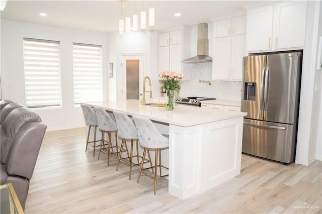 kitchen featuring pendant lighting, wall chimney range hood, stainless steel appliances, an island with sink, and white cabinets