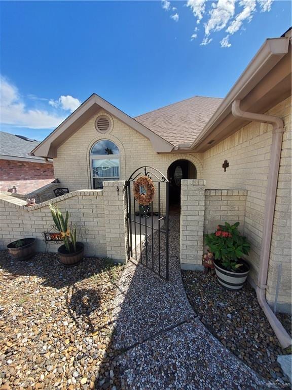 entrance to property featuring roof with shingles, a gate, fence, and brick siding