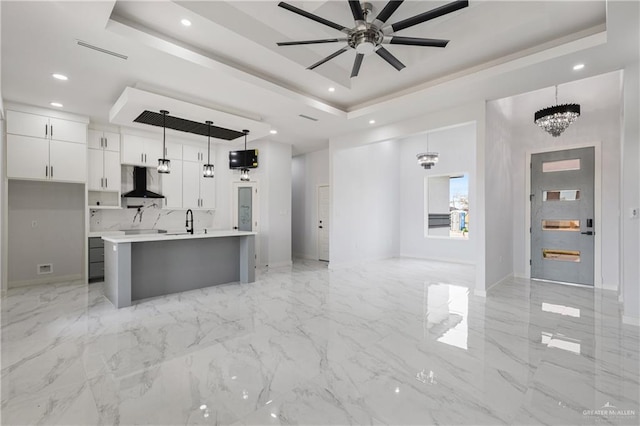 kitchen with pendant lighting, a kitchen island with sink, white cabinets, a raised ceiling, and wall chimney exhaust hood