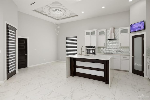 kitchen featuring wall chimney range hood, sink, white cabinetry, a center island with sink, and oven