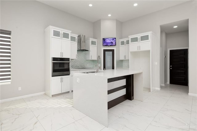 kitchen with white cabinets, wall chimney exhaust hood, stainless steel oven, and a center island with sink