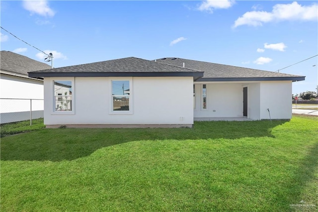 rear view of house featuring roof with shingles, fence, a lawn, and stucco siding