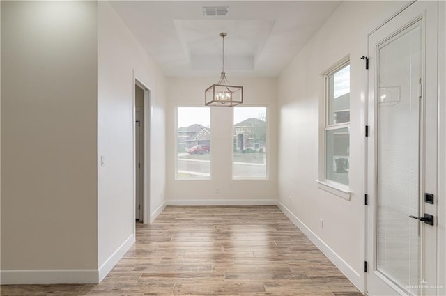 unfurnished dining area featuring baseboards, visible vents, an inviting chandelier, a raised ceiling, and light wood-type flooring