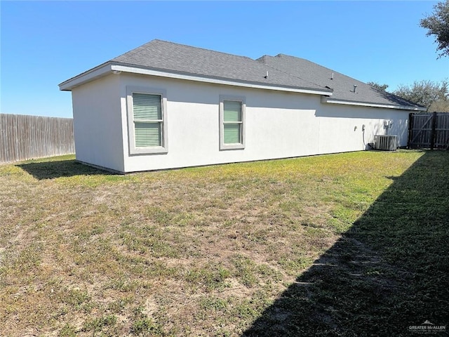 back of house with a shingled roof, central AC unit, fence, a yard, and stucco siding