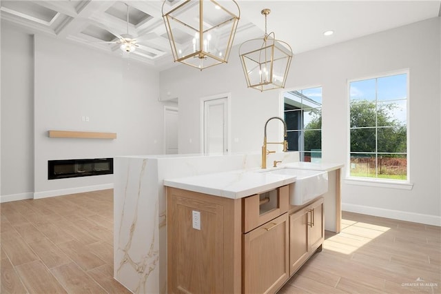 kitchen with coffered ceiling, sink, hanging light fixtures, light wood-type flooring, and an island with sink