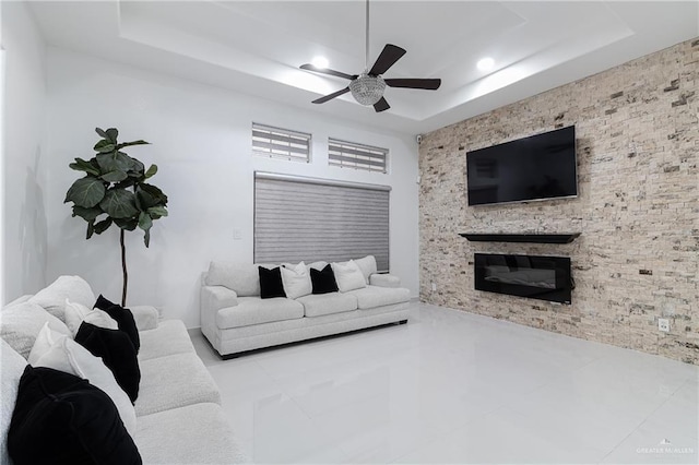 living room featuring a raised ceiling, a stone fireplace, and ceiling fan