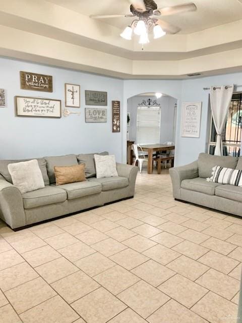 living area with arched walkways, light tile patterned floors, a ceiling fan, and a tray ceiling