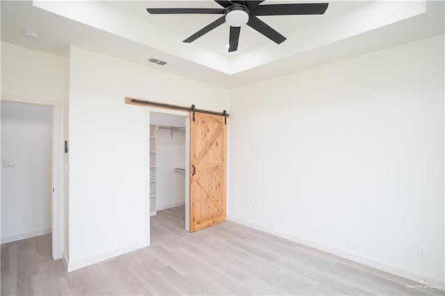 unfurnished bedroom with a tray ceiling, light hardwood / wood-style floors, a barn door, a walk in closet, and a closet