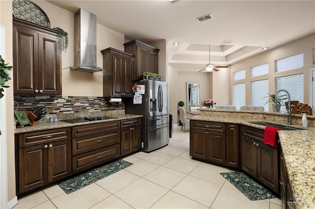 kitchen featuring stainless steel refrigerator with ice dispenser, sink, black electric cooktop, a tray ceiling, and wall chimney range hood