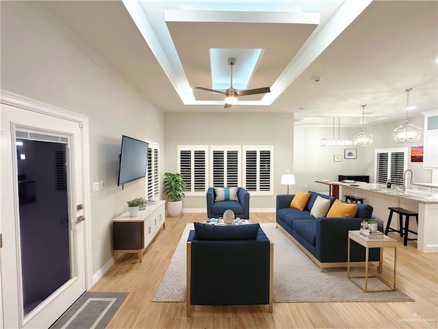 living room featuring ceiling fan with notable chandelier, light wood-type flooring, a raised ceiling, and sink