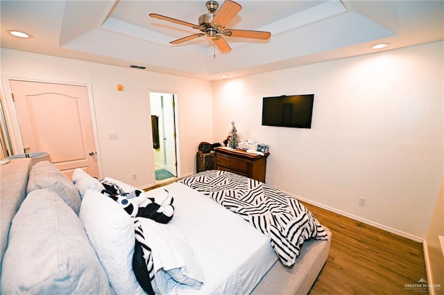 bedroom with wood-type flooring, a tray ceiling, ensuite bath, and ceiling fan