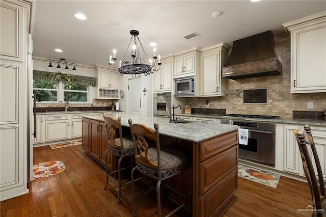 kitchen featuring appliances with stainless steel finishes, premium range hood, light stone counters, a center island with sink, and decorative light fixtures