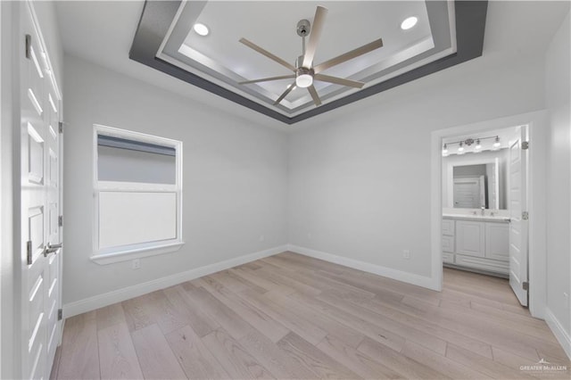 unfurnished bedroom with ceiling fan, sink, a tray ceiling, light wood-type flooring, and connected bathroom