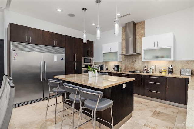 kitchen featuring wall chimney exhaust hood, built in appliances, decorative light fixtures, white cabinets, and a kitchen island