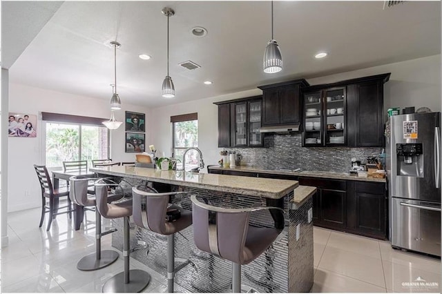 kitchen with decorative backsplash, a center island with sink, hanging light fixtures, and stainless steel refrigerator with ice dispenser