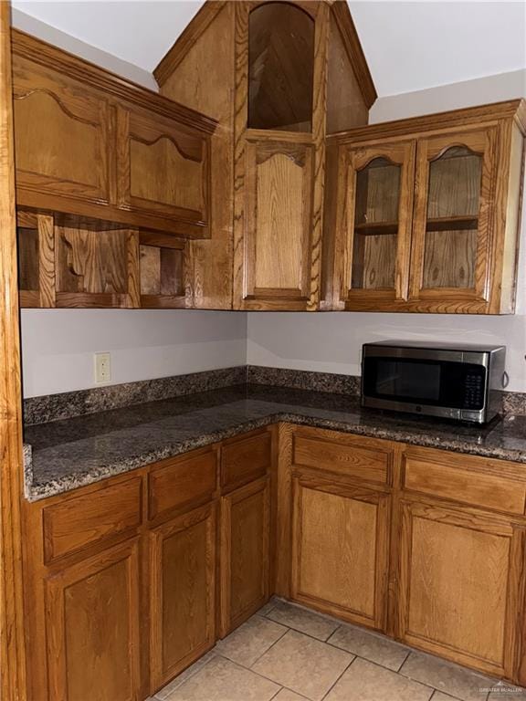 kitchen with brown cabinetry, glass insert cabinets, stainless steel microwave, dark stone countertops, and light tile patterned floors