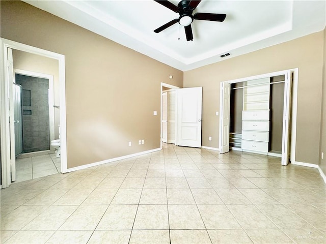 unfurnished bedroom with light tile patterned floors, a closet, a tray ceiling, ensuite bathroom, and ceiling fan