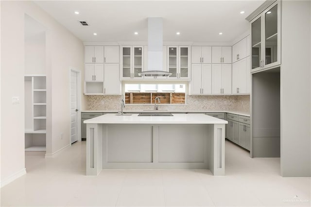 kitchen with sink, range hood, an island with sink, white cabinets, and decorative backsplash