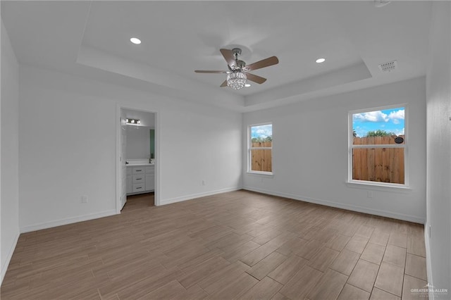 unfurnished bedroom featuring light wood-type flooring, ensuite bathroom, ceiling fan, and a tray ceiling