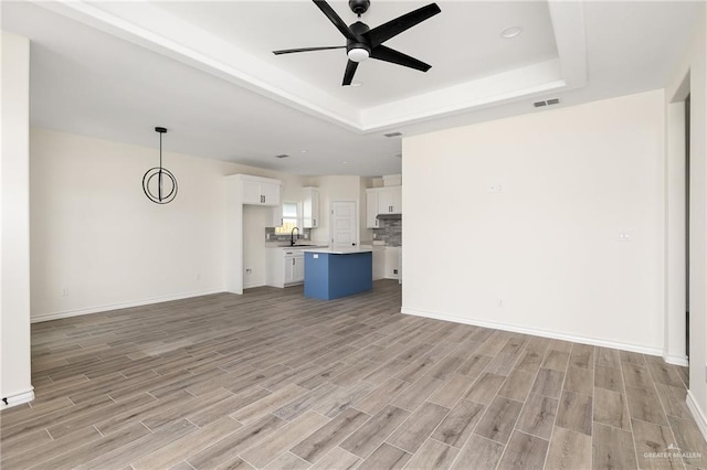 unfurnished living room with a raised ceiling, ceiling fan, sink, and light wood-type flooring