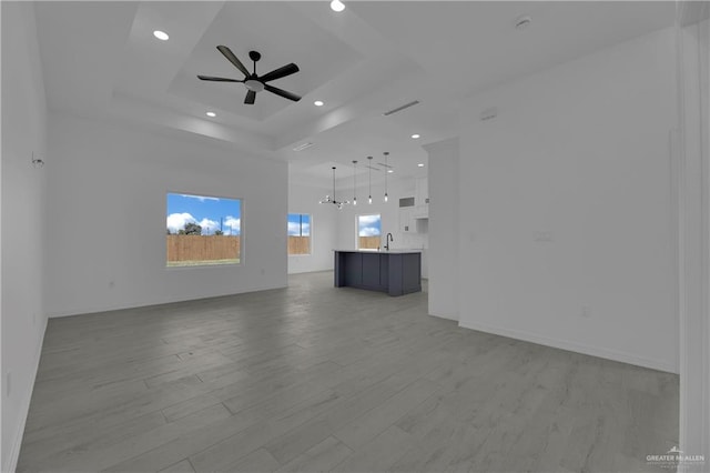 unfurnished living room with ceiling fan with notable chandelier, a raised ceiling, sink, a high ceiling, and light hardwood / wood-style floors