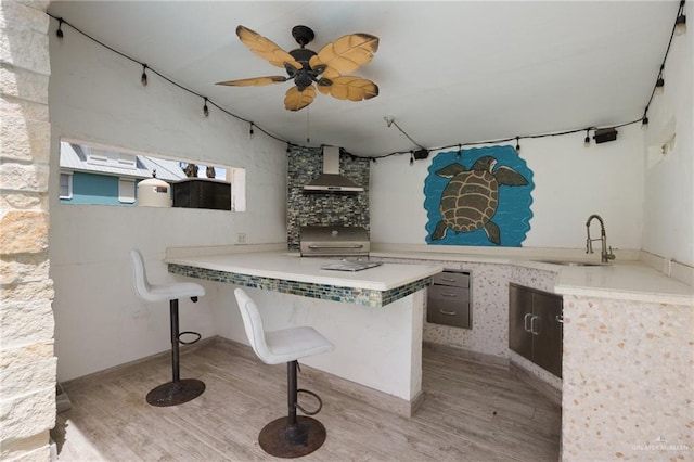 kitchen featuring ceiling fan, sink, wall chimney exhaust hood, a kitchen breakfast bar, and light wood-type flooring