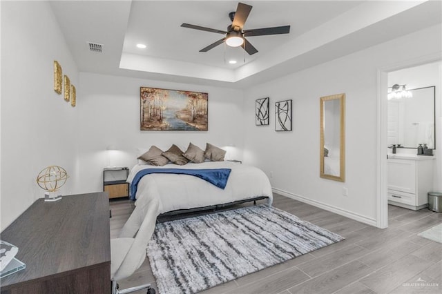 bedroom with ceiling fan, ensuite bathroom, a raised ceiling, and light hardwood / wood-style flooring