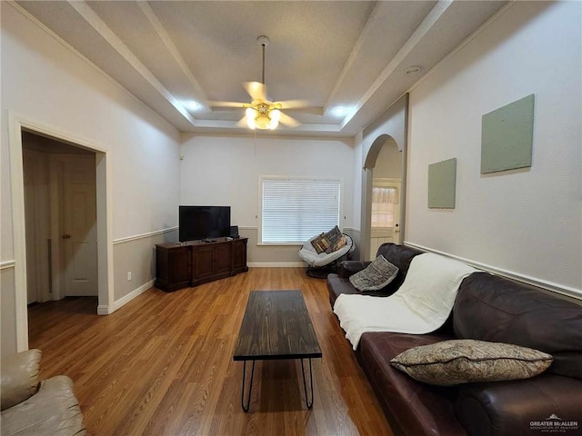 living area featuring baseboards, arched walkways, ceiling fan, a raised ceiling, and light wood-type flooring