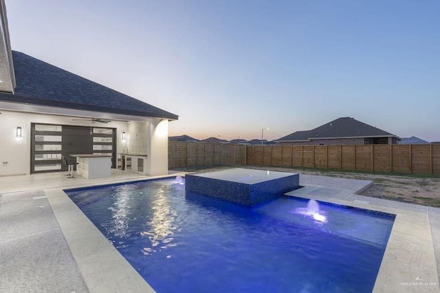 pool at dusk with an in ground hot tub, pool water feature, ceiling fan, and a patio