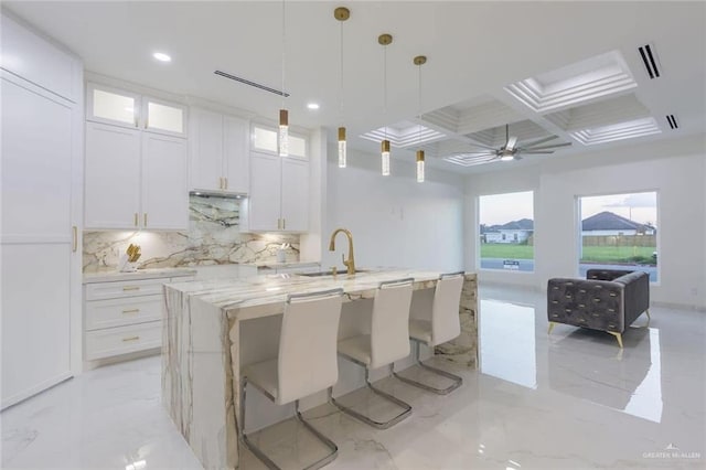 kitchen featuring coffered ceiling, white cabinetry, light stone counters, decorative light fixtures, and a center island with sink