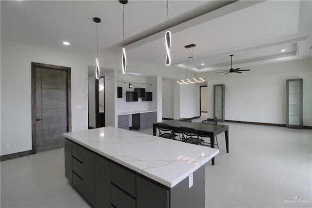 kitchen with ceiling fan, light stone countertops, hanging light fixtures, a spacious island, and a tray ceiling