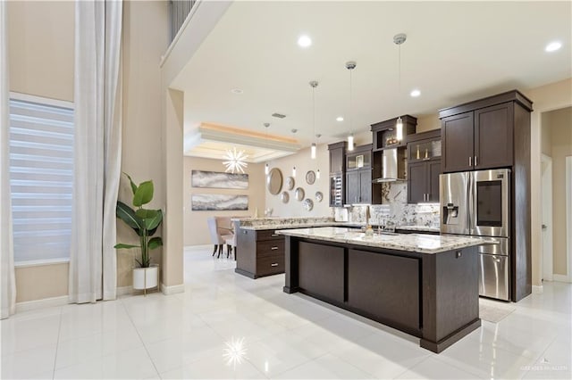 kitchen featuring dark brown cabinetry, light stone countertops, wall chimney exhaust hood, stainless steel fridge, and an island with sink