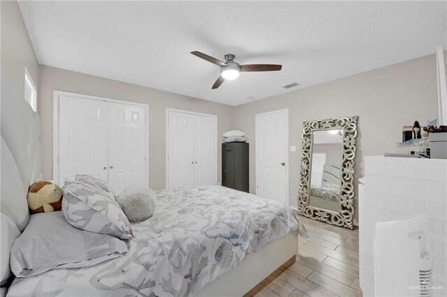 bedroom with multiple closets, visible vents, light wood-style floors, a ceiling fan, and a textured ceiling