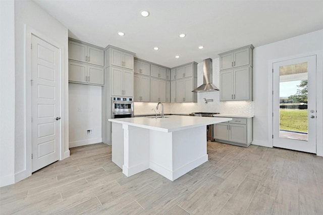 kitchen with stainless steel double oven, sink, an island with sink, wall chimney range hood, and gray cabinets