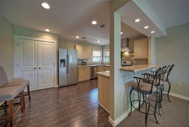 kitchen with light brown cabinetry, kitchen peninsula, appliances with stainless steel finishes, wall chimney exhaust hood, and a kitchen bar