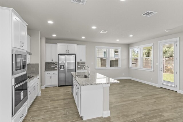 kitchen with an island with sink, sink, white cabinetry, stainless steel appliances, and light wood-type flooring