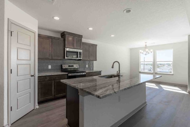 kitchen featuring a kitchen island with sink, sink, light stone counters, a chandelier, and stainless steel appliances