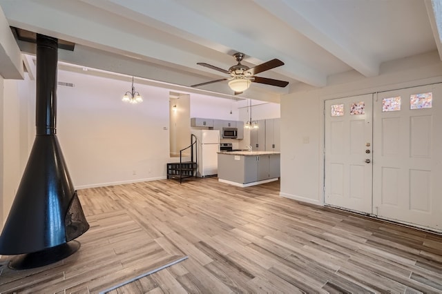 unfurnished living room featuring beam ceiling, ceiling fan with notable chandelier, a wood stove, and light wood-type flooring