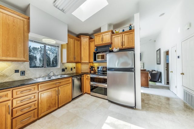 kitchen featuring a skylight, sink, backsplash, dark stone counters, and appliances with stainless steel finishes