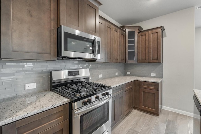 kitchen featuring appliances with stainless steel finishes, backsplash, dark brown cabinets, light stone counters, and light wood-type flooring