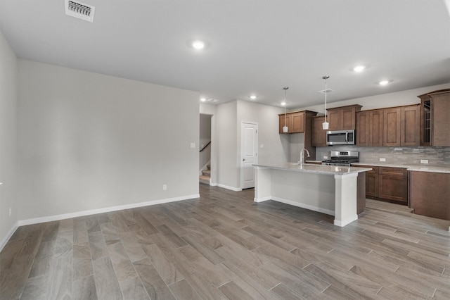 kitchen featuring appliances with stainless steel finishes, light wood-type flooring, tasteful backsplash, light stone countertops, and a kitchen island with sink