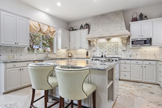 kitchen featuring stainless steel microwave, a kitchen island with sink, custom range hood, and white cabinetry